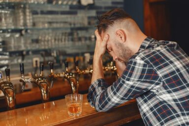 Opitý muž sedí pri bare s hlavou v rukách, vedľa seba má na bare položený pohár s hnedým alkoholom.